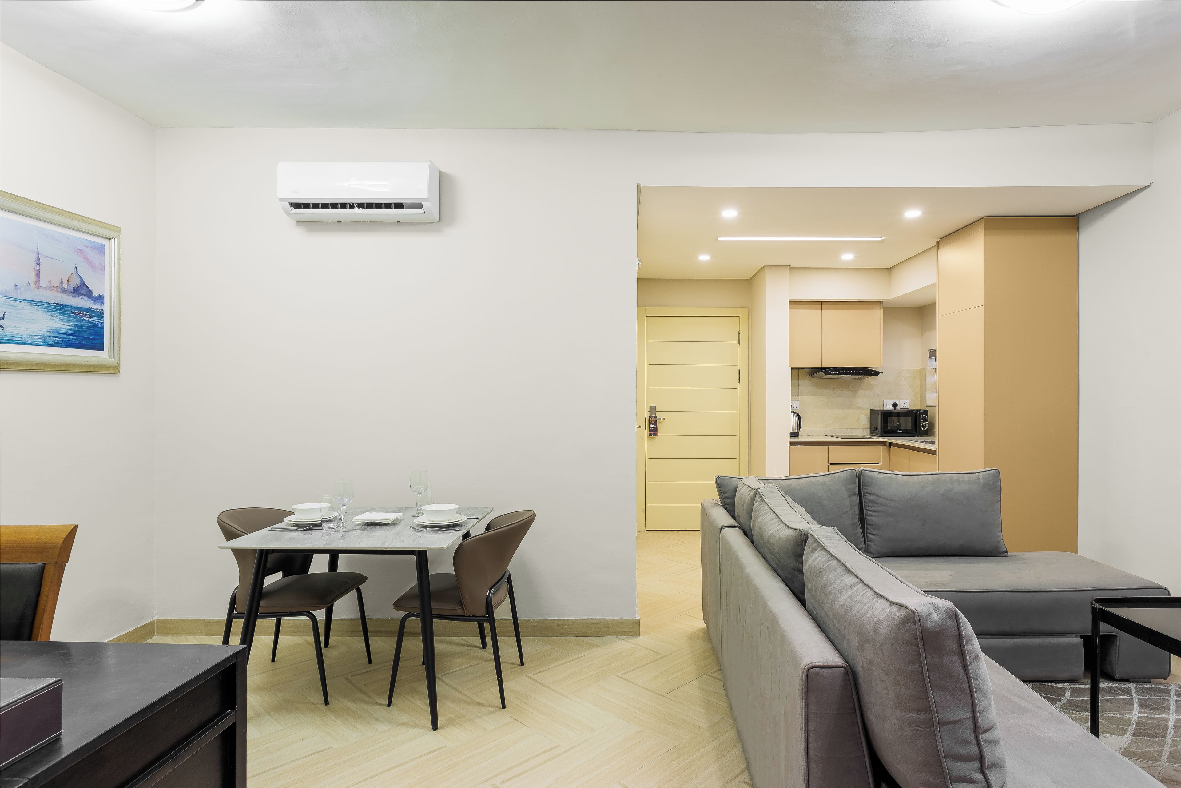 A modern living room with a sofa, dining table, and chairs. There is a kitchen area with cabinets and a microwave. An air conditioning unit is mounted on the wall.