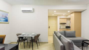 A modern living room with a sofa, dining table, and chairs. There is a kitchen area with cabinets and a microwave. An air conditioning unit is mounted on the wall.