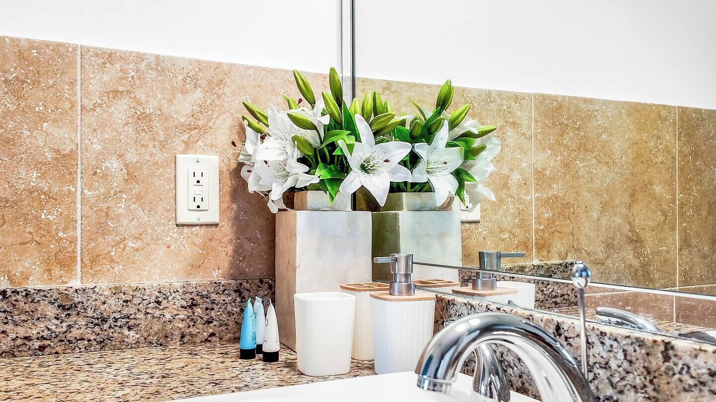 Un baño con encimera de granito, lavamanos con grifo moderno, espejo y planta en maceta con flores blancas.