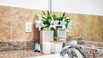 Un baño con encimera de granito, lavamanos con grifo moderno, espejo y planta en maceta con flores blancas.