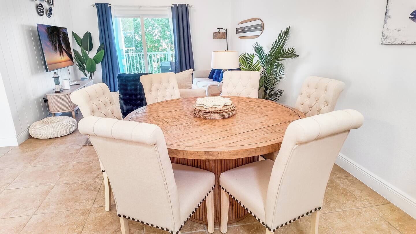 A round wooden dining table with six upholstered chairs, a woven centerpiece, and a television on a stand in the background.