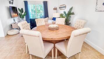 A round wooden dining table with six upholstered chairs, a woven centerpiece, and a television on a stand in the background.