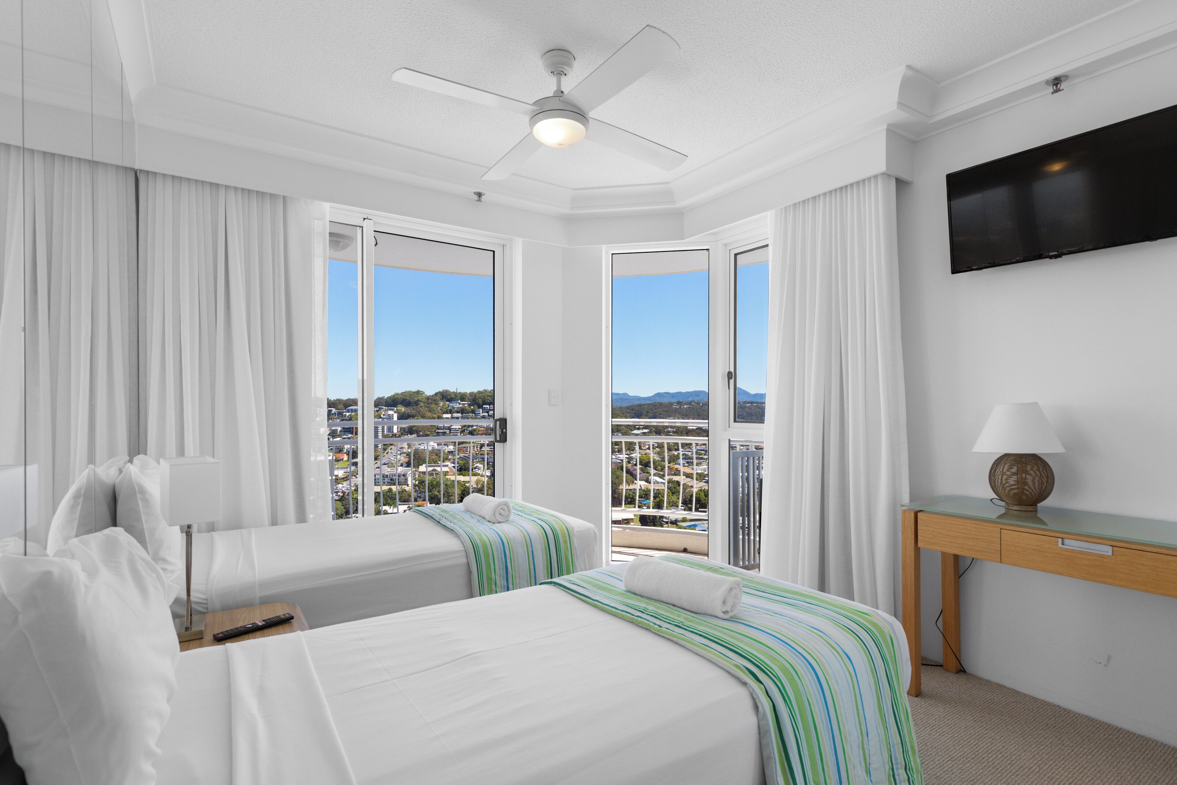 A hotel room with two beds, a ceiling fan, a TV, and a view of the city.