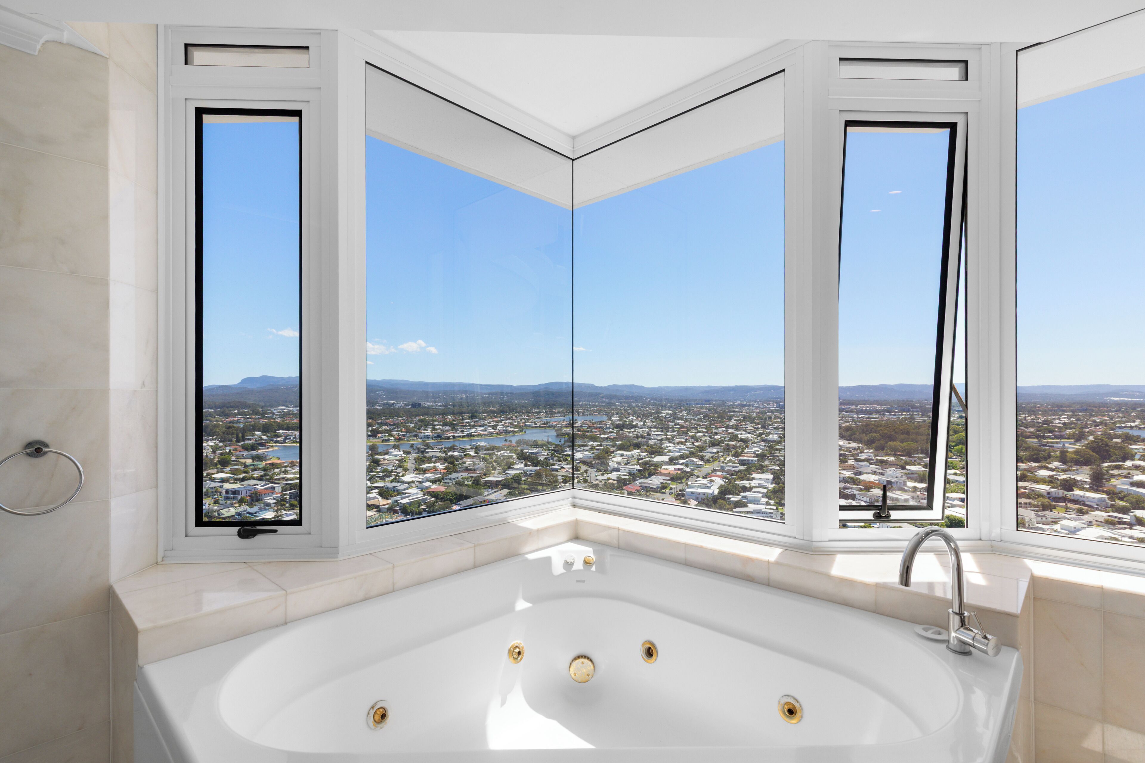 A modern bathroom with a large bathtub, three windows offering a city view, and a visible shower area.