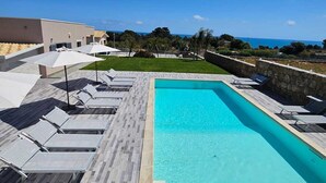 Una piscina rectangular con borde de piedra, rodeada de sillas de descanso y sombrillas, con vista despejada al océano y una zona de césped al fondo.