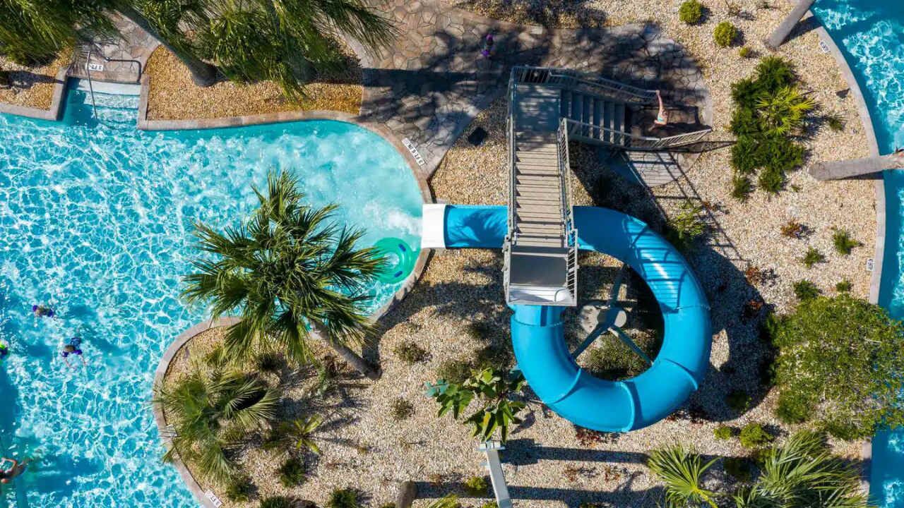 Vue aérienne d’une piscine avec un toboggan, entourée de palmiers et bordée d’une plage de galets.