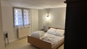 Ein Schlafzimmer mit einem hölzernen Bett, einem Nachttisch mit Lampe, einem Fenster mit Spitzenvorhängen und einer Heizung.