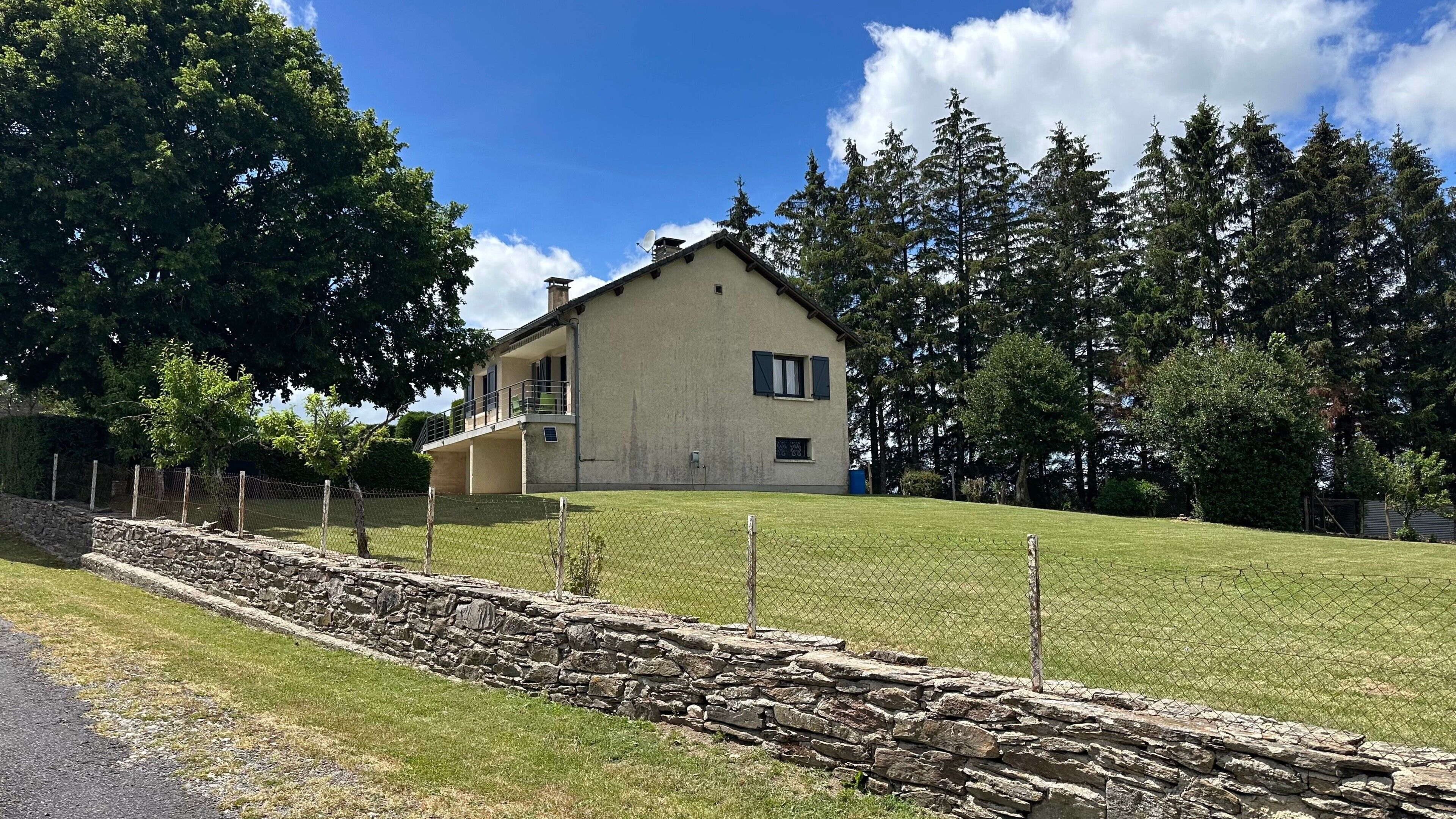 Ein zweistöckiges Haus mit einer Steinmauer im Vordergrund, umgeben von Bäumen und blauem Himmel.