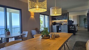 A modern kitchen with a central island, a dining area with a wooden table, and large windows offering a view of the outdoors. - Casa do Mundo Madeira (Santa Cruz)