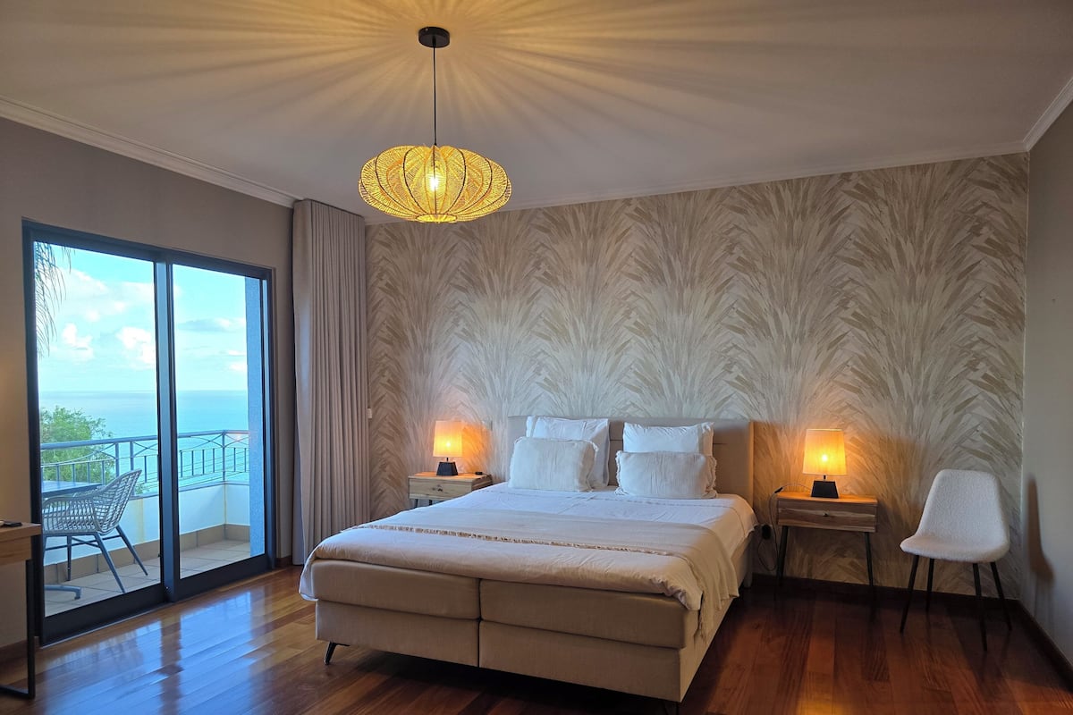 Un dormitorio con una cama grande, dos mesitas de noche con lámparas, vista al mar a través de un ventanal corredero y un papel tapiz estampado.