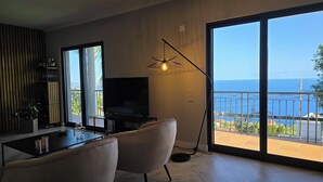 A modern living room with a large TV, two armchairs, a dining table, and a view of the sea through a balcony. - Casa do Mundo Madeira (Santa Cruz)