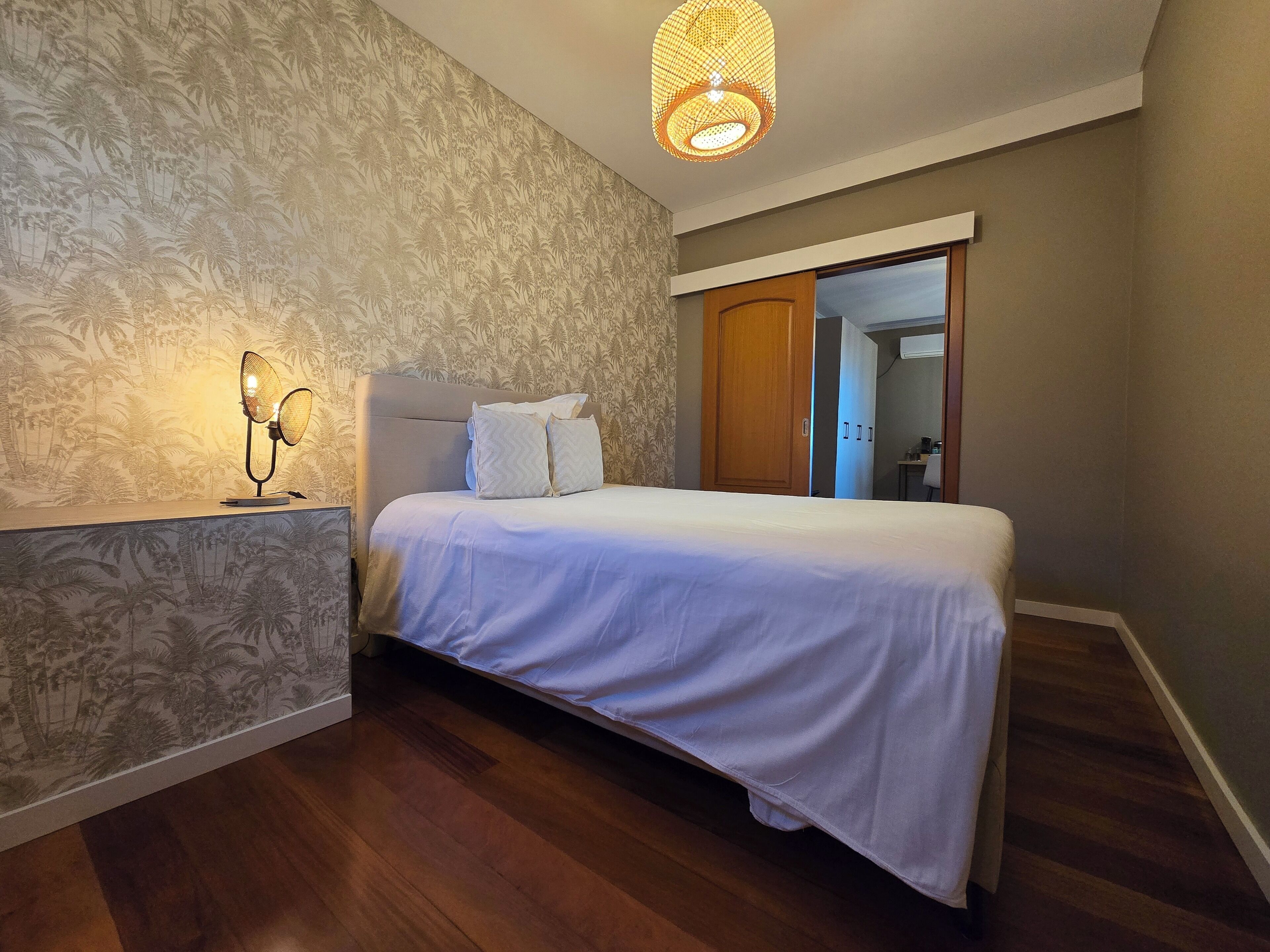 A bedroom with a bed, a nightstand, a wall-mounted lamp, and a door leading to a bathroom.