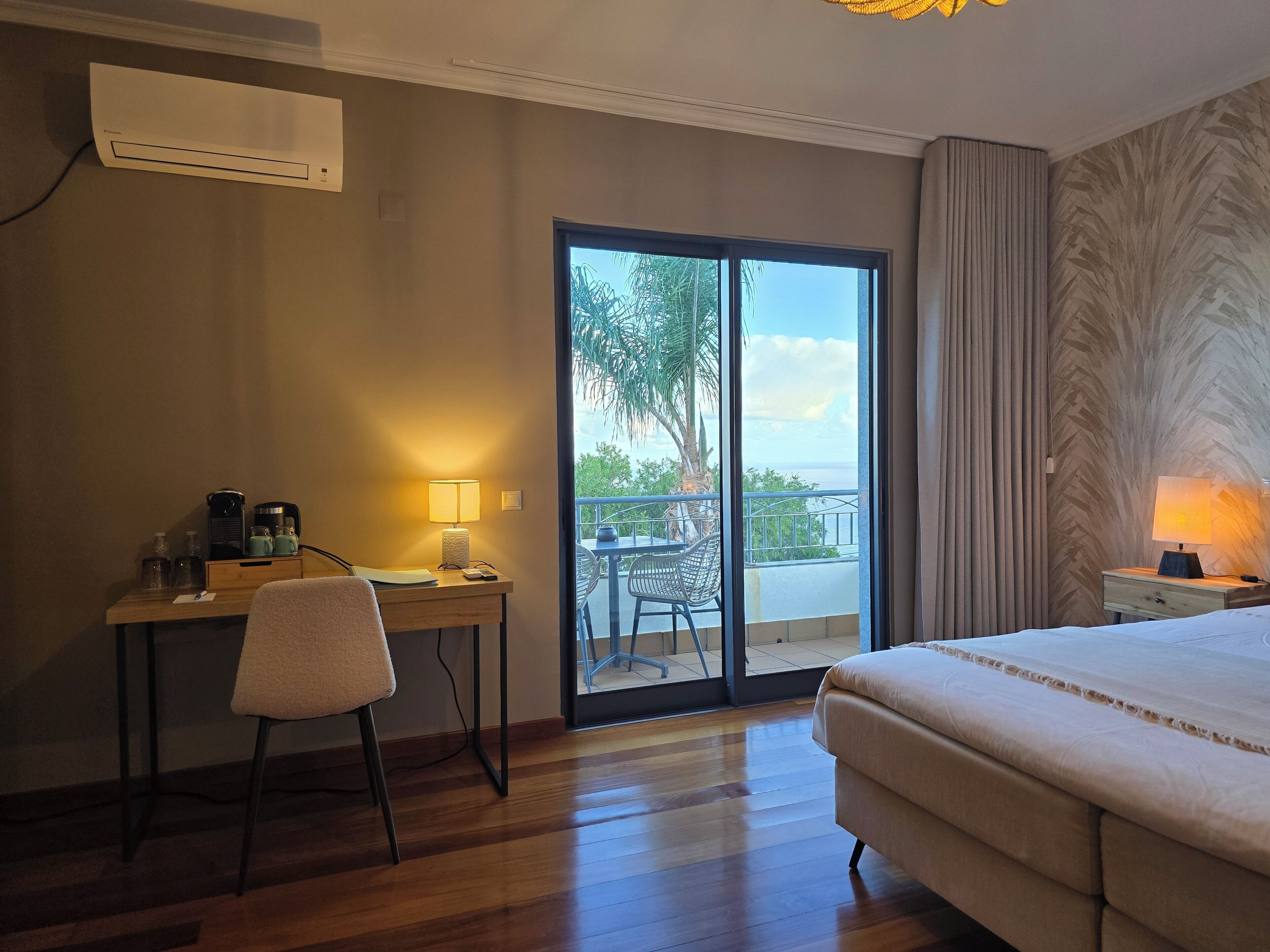 A hotel room with a bed, a desk with a chair, a lamp, and a view of the outdoors through a sliding door.