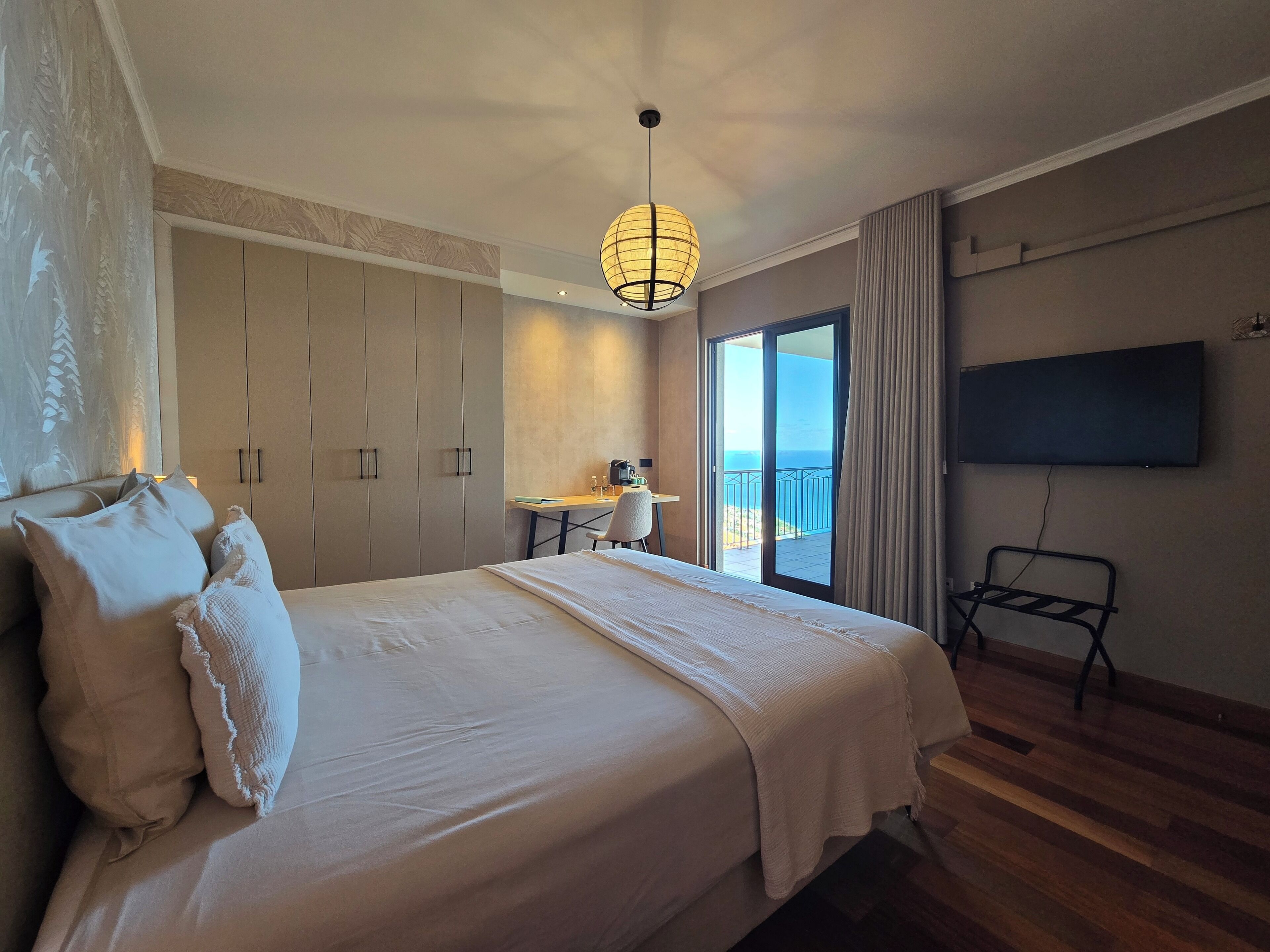 A hotel room with a large bed, a desk, a chair, a TV, and a balcony with a view of the sea.