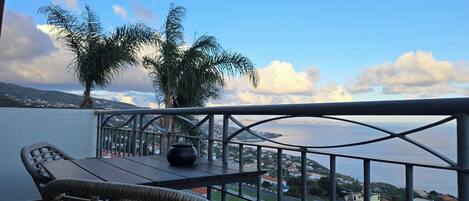 A balcony with a wicker chair, a table, and a vase. It overlooks a body of water with a distant shoreline and palm trees.