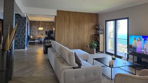 A modern living room with a sofa, a coffee table, and a TV. There is a wooden panel wall and a view of the outdoors. - Casa do Mundo Madeira (Santa Cruz)