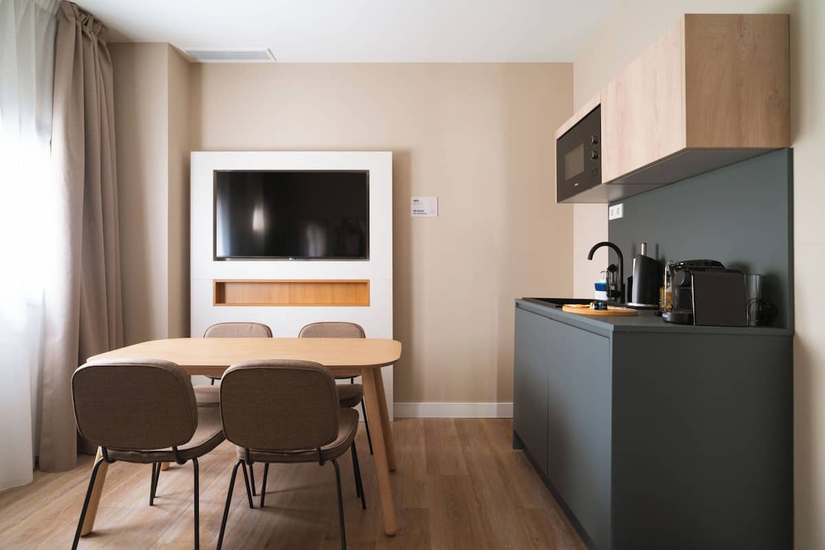 A compact kitchen with a microwave, toaster, and coffee maker. There is a dining table with four chairs and a large flat-screen TV mounted on the wall.