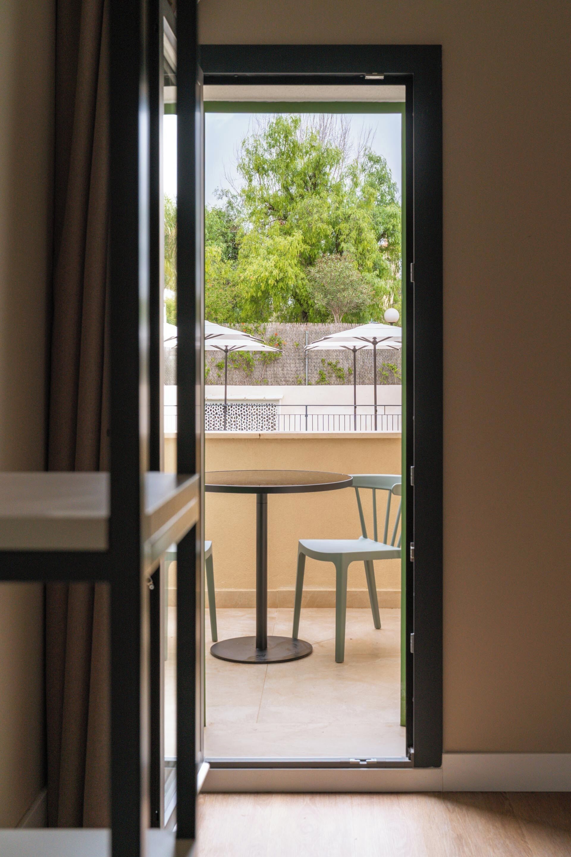 Puerta abierta que da a un patio exterior con una mesa y sillas, rodeado de follaje y sombrillas.