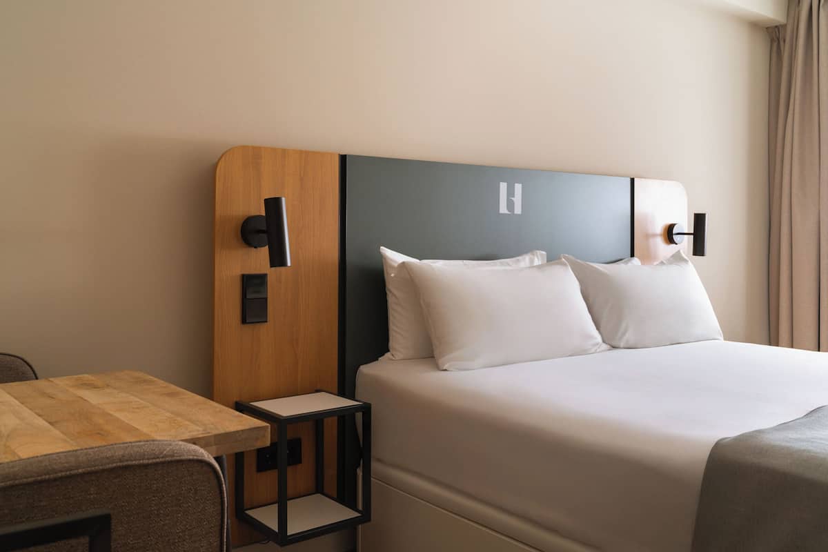 A hotel room with a wooden headboard, a small bedside table, a bed with white linens, and a view of the room's interior.
