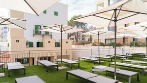 Un área de estar al aire libre con mesas y sombrillas, rodeada por un edificio blanco con balcones.