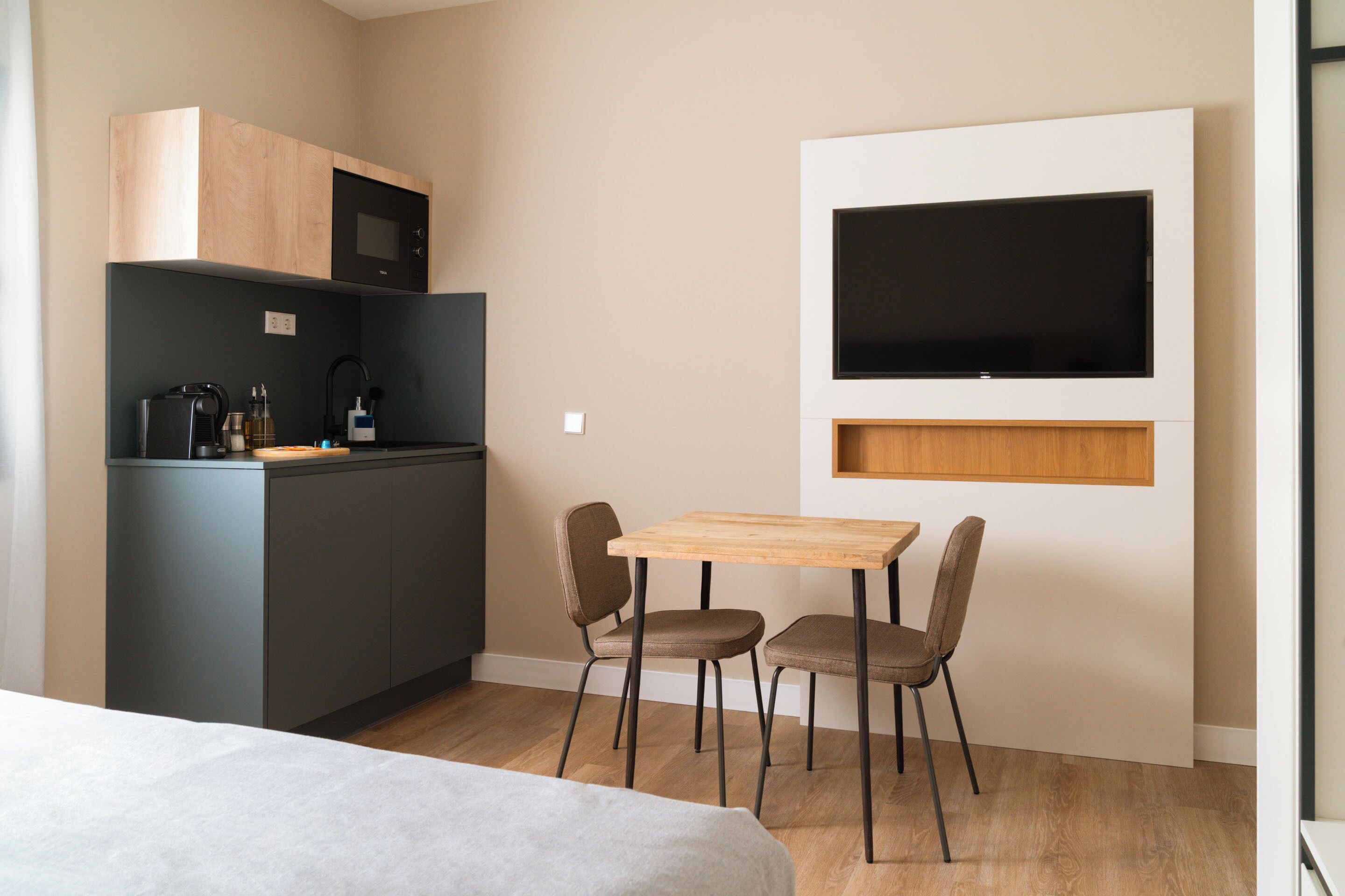 A compact kitchenette with a microwave, coffee maker, and small fridge. There is a wooden table with two chairs and a TV mounted on the wall.