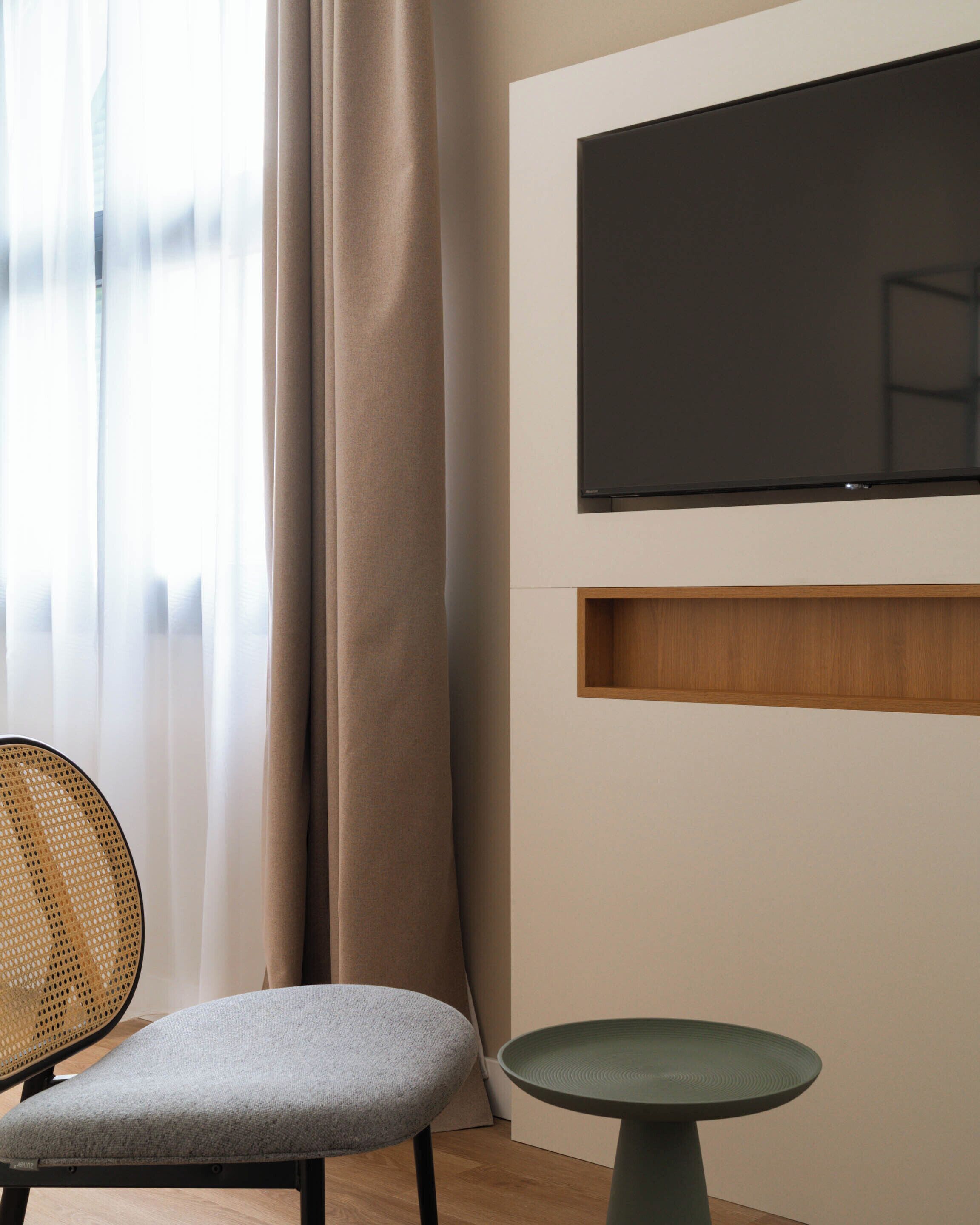 A modern room with a flat-screen TV mounted on the wall, a grey cushioned chair, a round table, and a window with sheer curtains.