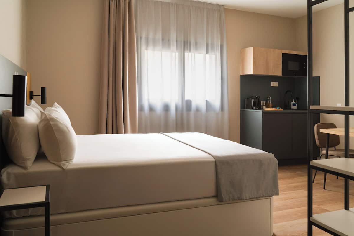 A modern hotel room with a bed, a desk, and a kitchenette.