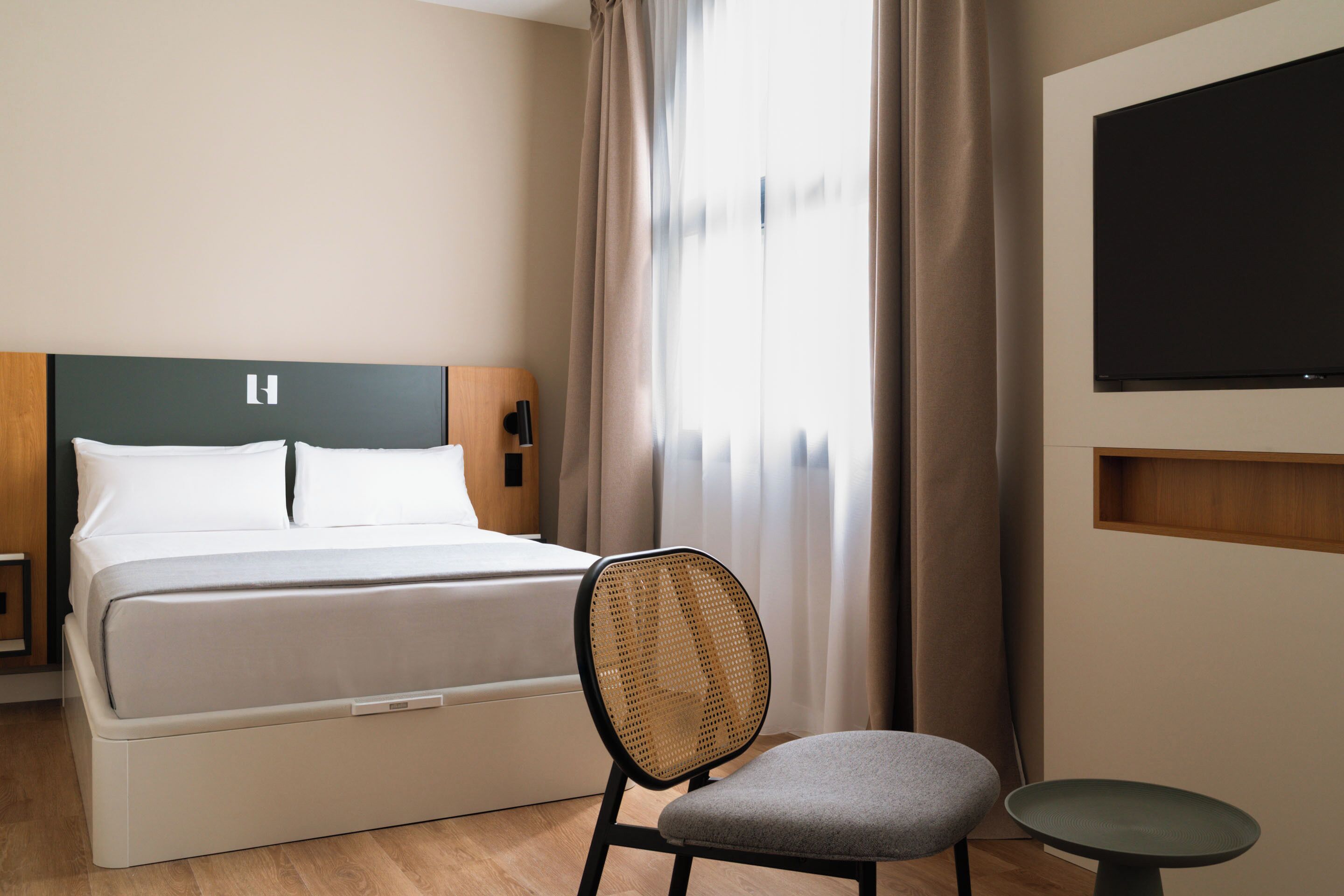 A hotel room with a bed, a chair, a small table, and a TV mounted on the wall.