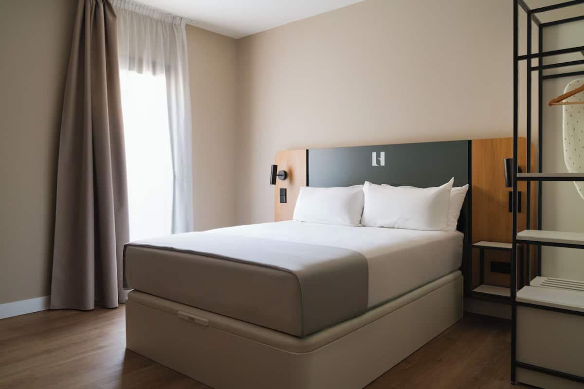 A modern hotel room with a large bed, a wooden headboard, a bedside table, and a wardrobe.