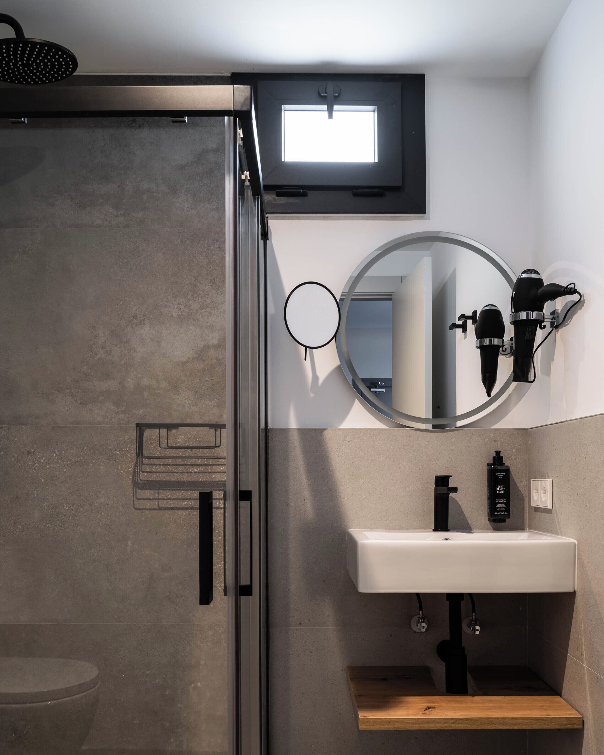 Un baño moderno con mampara de vidrio, espejo redondo, secador de pelo negro y lavamanos blanco.
