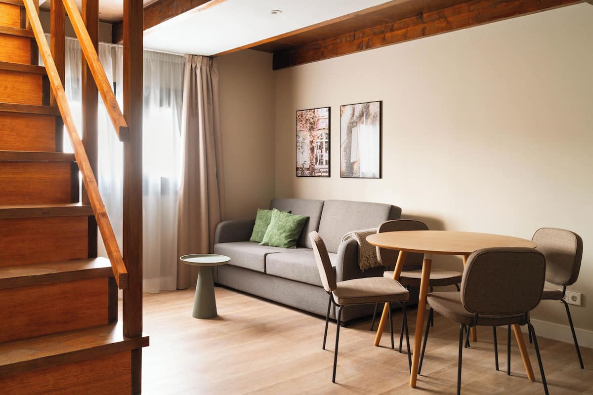 A modern living room with a wooden staircase, a grey sofa, a round wooden table, and two framed pictures on the wall.