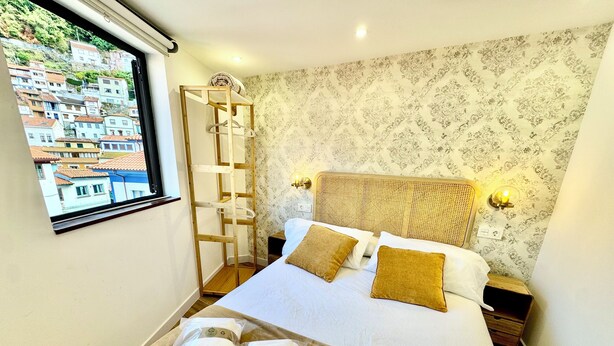 A hotel room with a bed, two pillows, a window with a city view, a wooden ladder, and a wall with patterned wallpaper. - Gestviva Casa Urbanin III (Cudillero)