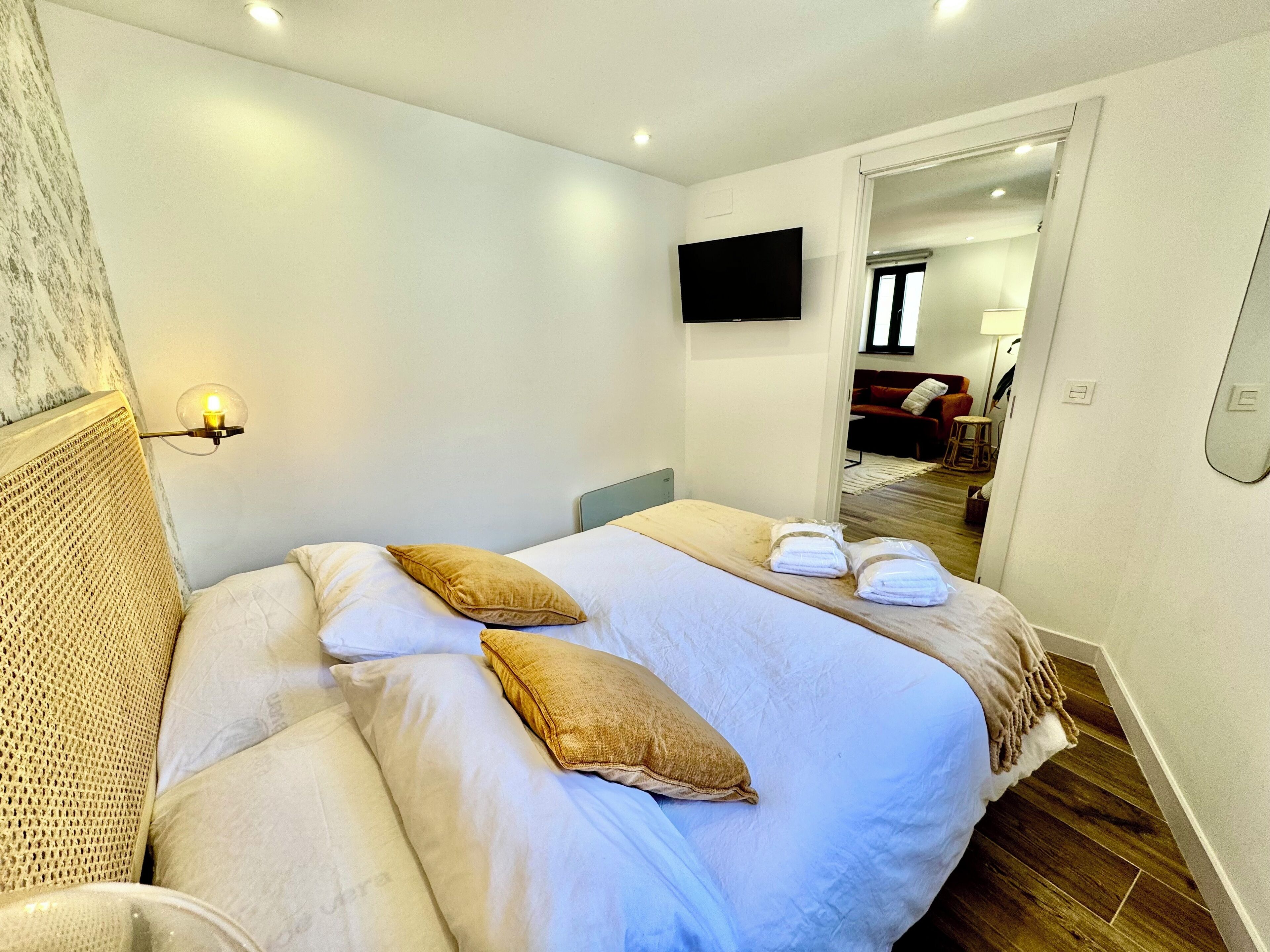 Een net opgemaakt bed met witte lakens en twee beige kussens, een televisie aan de muur en een houten vloer.