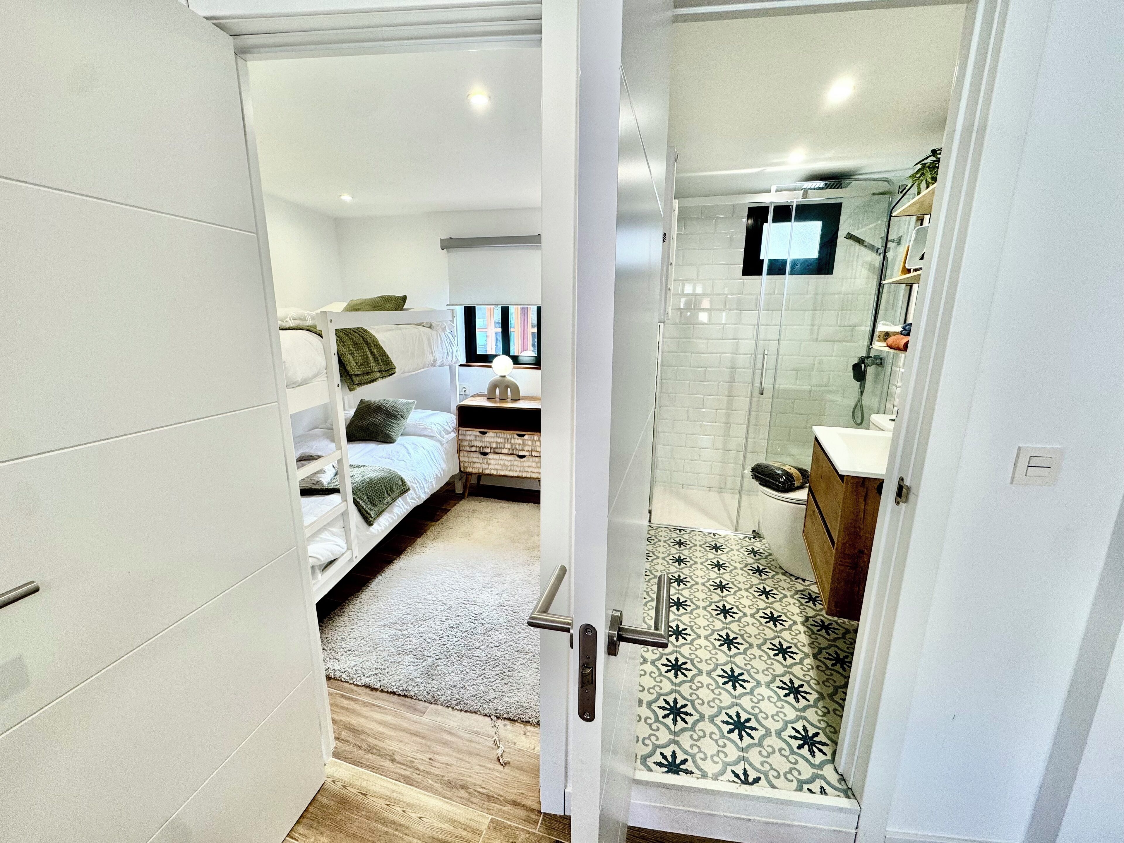 Un dormitorio moderno y funcional con literas, una mesita de noche pequeña y una zona de ducha con puerta de cristal, con vistas al exterior a través de una ventana.