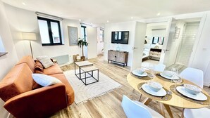A modern living room with a brown sofa, a coffee table, and a dining area with a round table set for four. - Gestviva Casa Urbanin III (Cudillero)