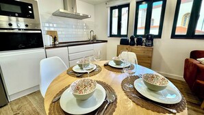 A modern kitchen with white cabinets, a stainless steel range hood, and a dining area with a wooden table set for two. - Gestviva Casa Urbanin III (Cudillero)
