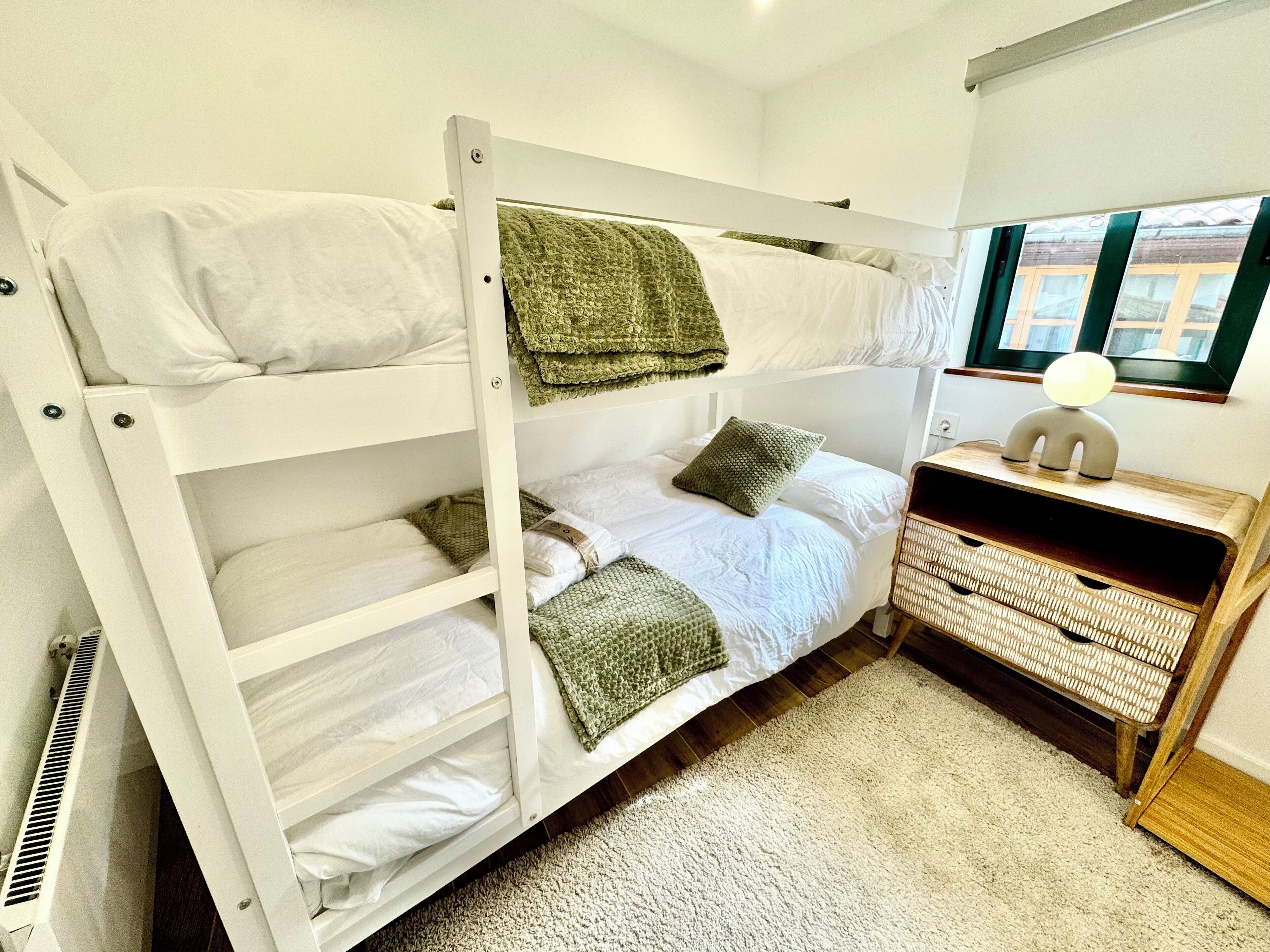 Una cama individual con ropa de cama blanca y mantas verdes, una pequeña mesita de madera con una lámpara, y una ventana con vistas a edificios.