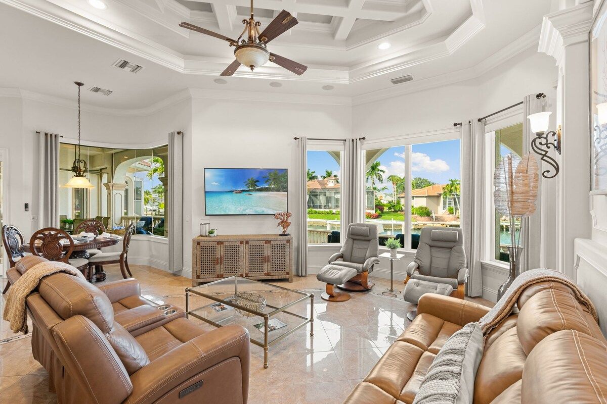 A spacious living room with a ceiling fan, leather sofas, a coffee table, and a view of a tropical landscape through large windows.