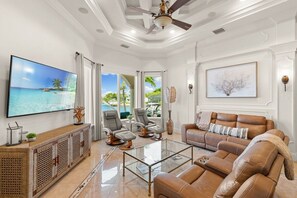 A spacious living room with a large flat-screen TV, leather sofas, a glass coffee table, and a ceiling fan.