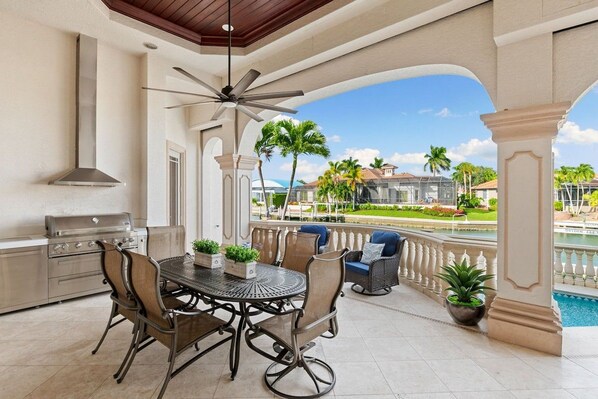 An outdoor patio with a grill, dining table, and chairs, overlooking a pool and a landscaped area.