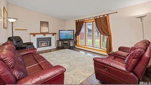 A living room with a fireplace, leather sofas, a television, and a large window with curtains. - Family 4BR Home With Patio by Willowcreek Park (Sparks)