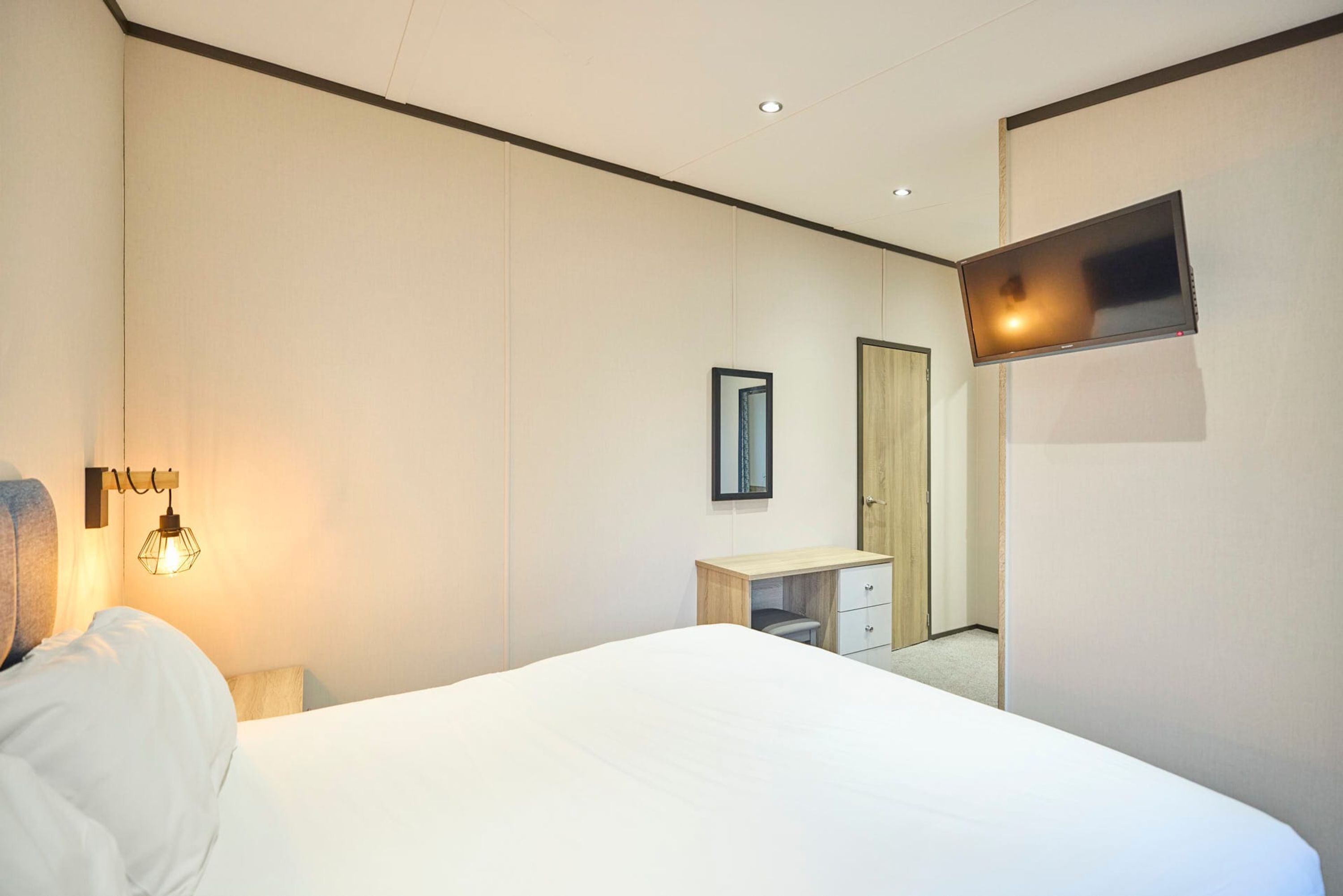 A hotel room with a bed, a desk, a mirror, and a flat-screen TV mounted on the wall.