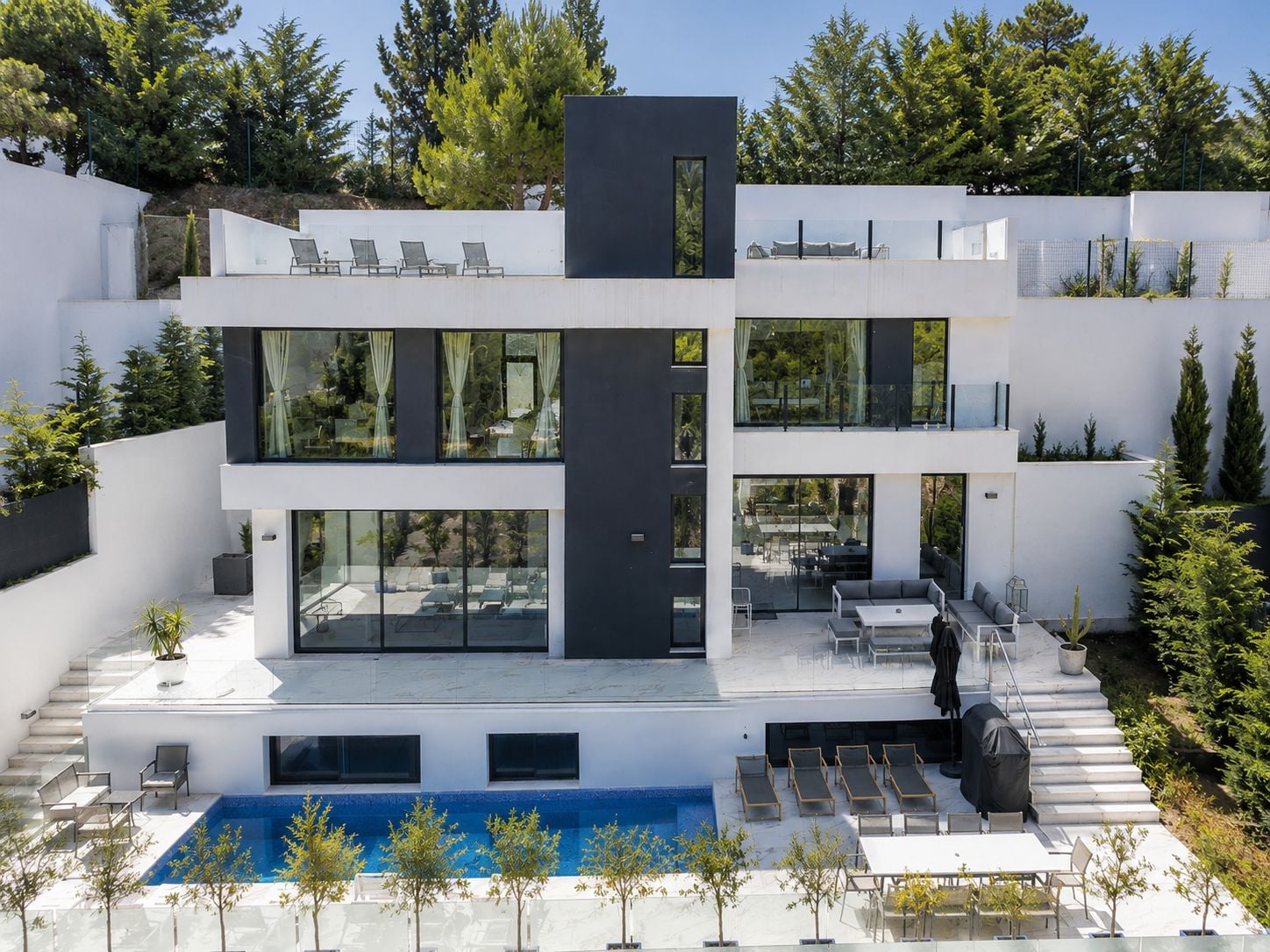A modern multi-story house with a pool, outdoor seating, and a view of trees.