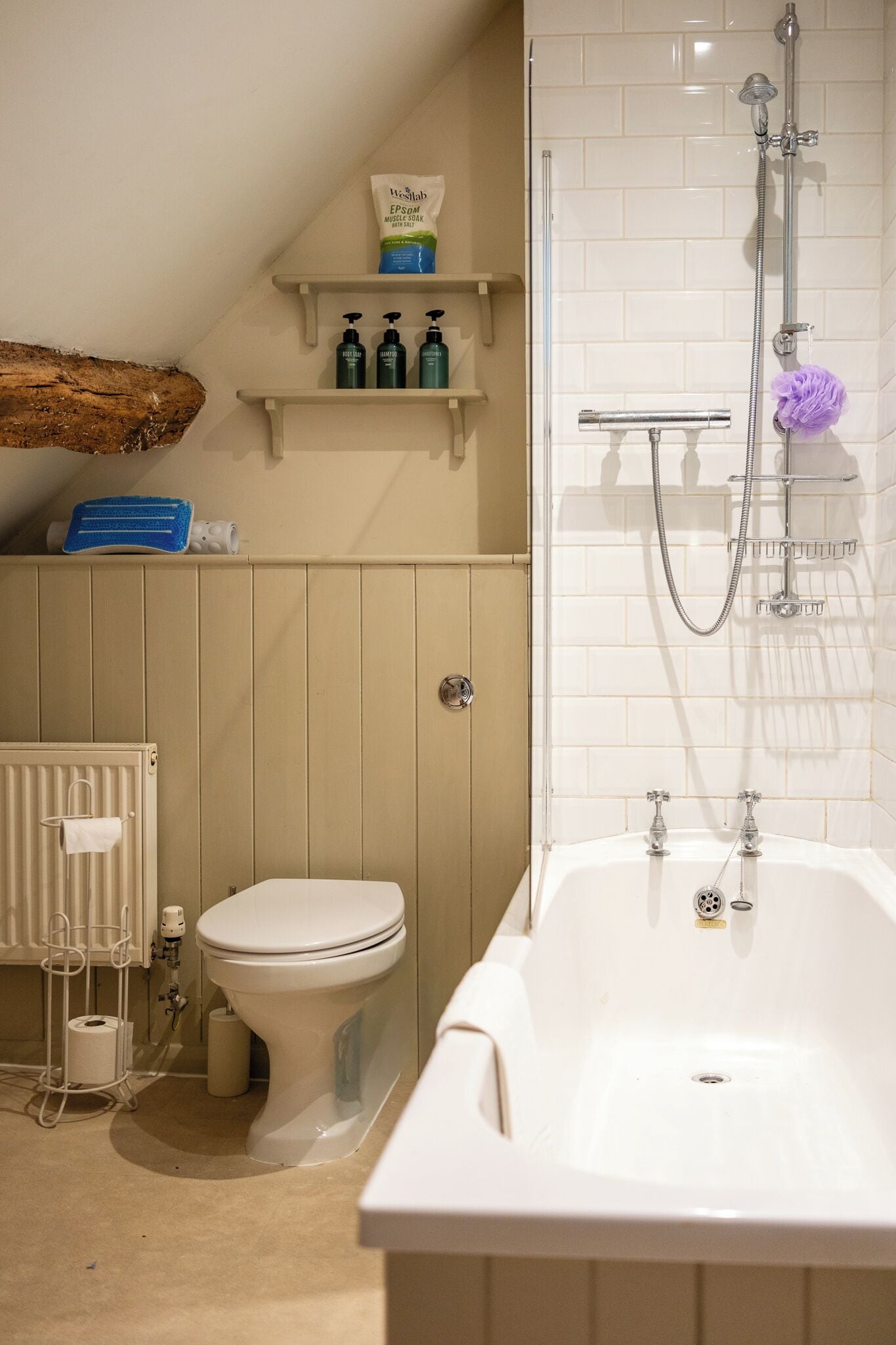Ein Badezimmer mit einer Badewanne, Toilette und einer Dusche. Es gibt Regale mit Pflegeprodukten und einen Holzbalken über der Toilette.