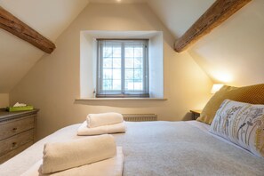 A bedroom with a sloped ceiling, a window with blinds, a bedside table with a lamp, a bed with white bedding, and a decorative pillow. - Beautiful Cottage in the Cotswolds (Chipping Campden)