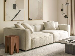 Una sala moderna con un sofá color beige, un taburete de madera y una mesa de centro. Hay pinturas abstractas en la pared y una lámpara de pie.