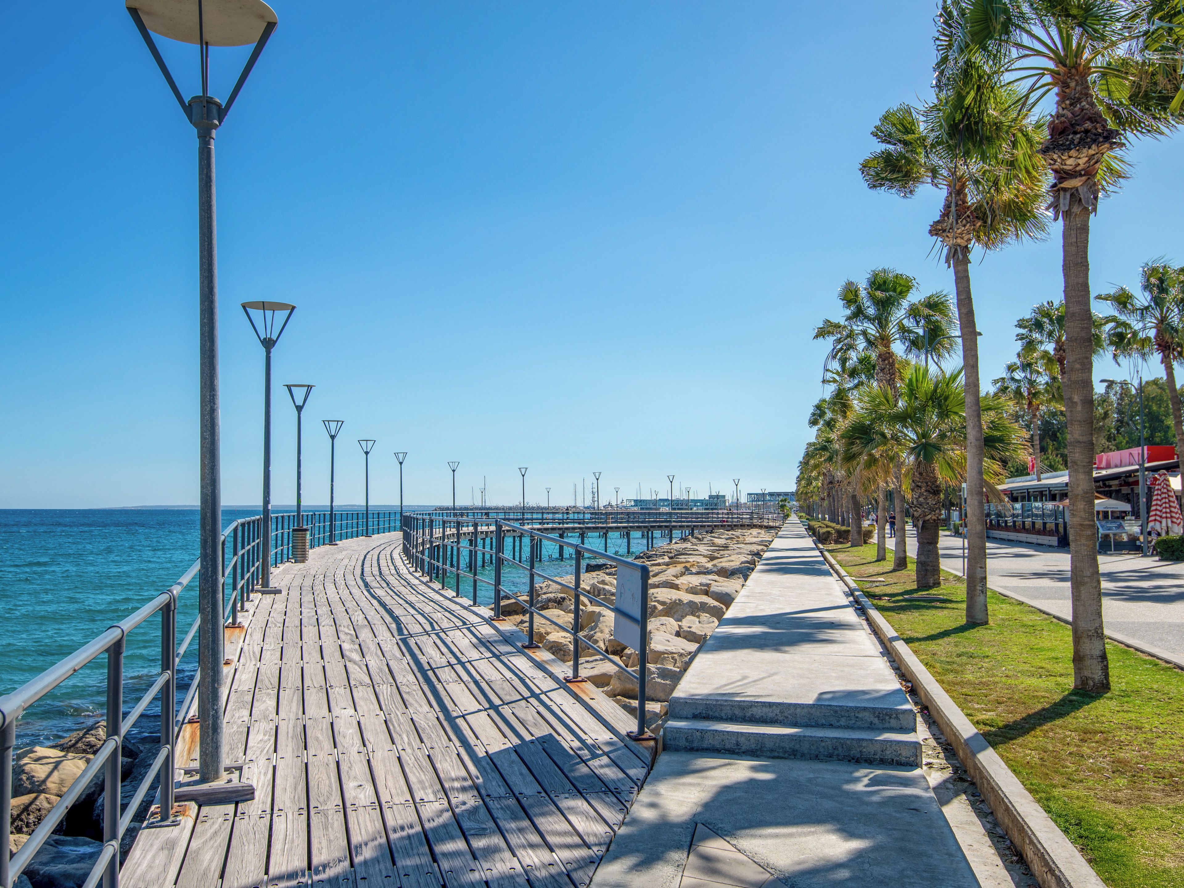 A wooden boardwalk along a coastal area with street lamps, palm trees, and a clear blue sky.