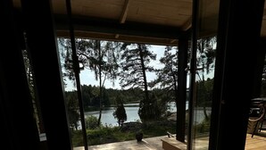 A view from inside a room with a glass door leading to an outdoor deck. The deck has a wooden floor and is surrounded by trees and a body of water.