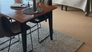 A hotel room with a wooden dining table, a vase with dried plants, a bed with a quilted bedspread, and a view of a lake through a window.