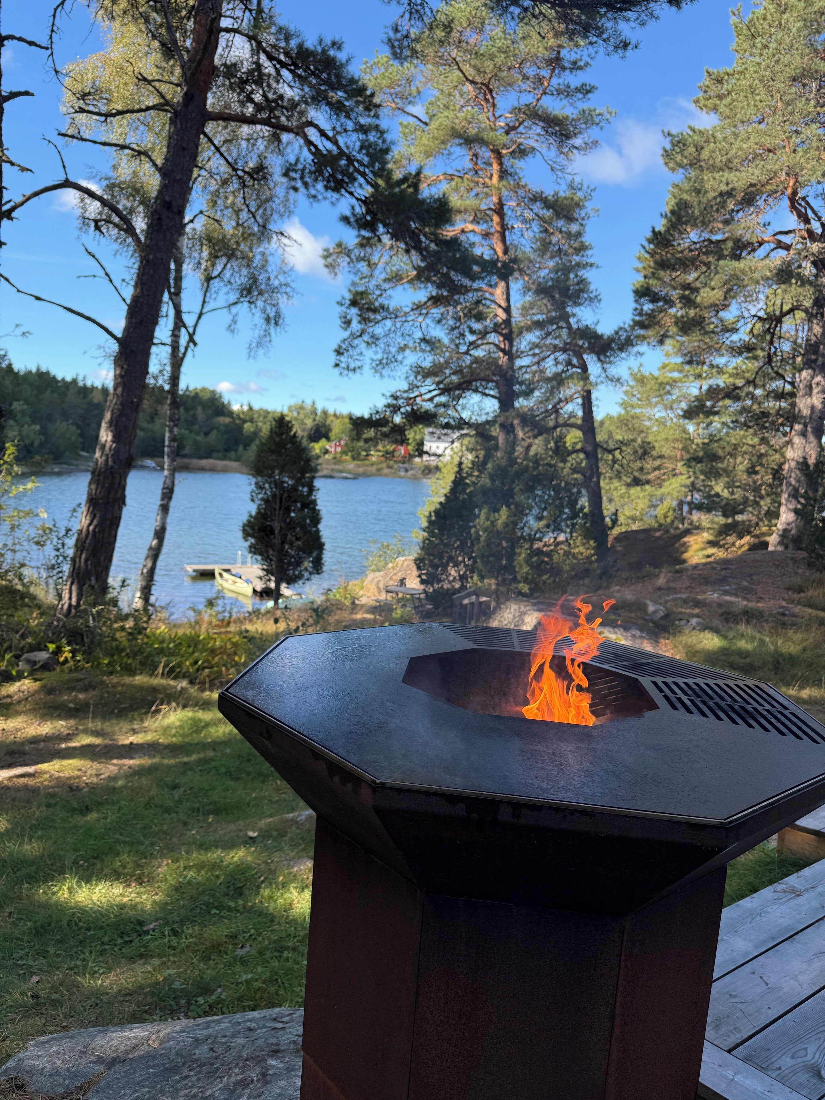 A hexagonal outdoor grill with a visible flame, surrounded by a natural landscape with trees and a lake.